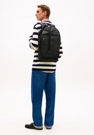 Μαύρο δερμάτινο σακίδιο με εμπρόσθια τσέπη με φερμουάρ, κλείσιμο με κορδόνι και ρυθμιζόμενους ιμάντες, φορεμένο από άτομο με ριγωτό πουλόβερ και μπλε τζιν.