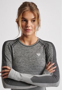 Ung kvinna med blont hår uppsatt, iklädd en grå och svart långärmad sporttröja, armarna korsade, tittar åt sidan.