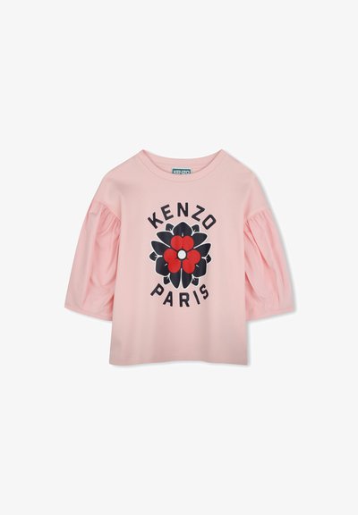 T-shirt rose à manches courtes avec des manches bouffantes, présentant un grand motif floral en rouge, noir et blanc, le texte "KENZO PARIS" en dessous de la conception.