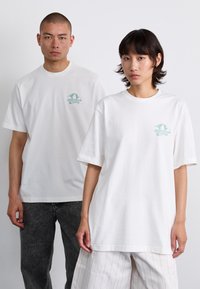 Białe bawełniane koszulki z małym turkusowym nadrukiem na piersi. Obie mają luźny krój; jedna zestawiona z szarymi dżinsami, druga z pasiastymi spodniami.