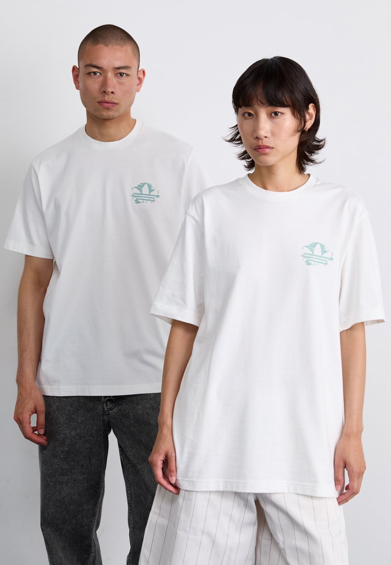 Witte katoenen T-shirts met een kleine groene afbeelding op de borst. Beide dragen een losse pasvorm; de ene gecombineerd met grijze jeans, de andere met gestreepte broek.