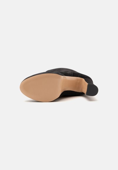 Chaussure en cuir noir posée sur le côté, montrant une semelle beige lisse et un petit talon sur un fond blanc uni.