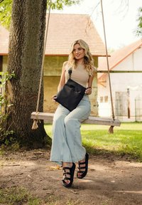 Čern�á kabelka přes rameno s jediným popruhem, hladkým povrchem a minimalistickým designem. Modelka má na sobě světle modré zvonové džíny a černé sandály s pásky.