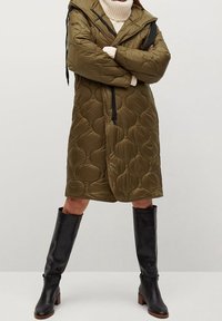 Olivengrøn quiltet frakke med hætte, der har et tekstureret, oppustet design og snoredetaljer, kombineret med knælange sorte læderstøvler.