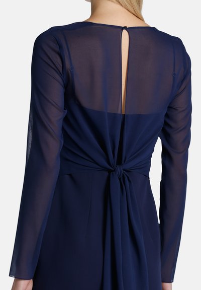 Tamsiai mėlyna suknelė su ilgomis permatomomis rankovėmis, mazguotu nugaros akcentu ir raktelio formos iškirpte nugaroje. Lygus audinys su plazdančiu dizainu.
