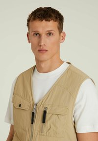 Beež kerge vest kahe lukuga tasku ja nupuga taskuga. Kanda valge ümaralise kaelusega särke peal. Sile tekstuur, minimaalne disain.