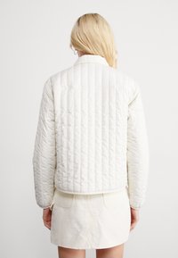 Valkoinen tikattu takki, jossa lyhyt leikkaus, kärkipäinen kaulus ja resorireunus. Sileä pinta vaakasuorilla ommelkuvioilla.