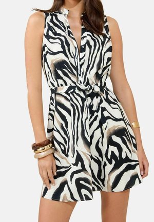 Femme portant une robe sans manches à imprimé zèbre avec ceinture à nouer, accessoirisée de plusieurs bracelets au poignet gauche et d’une manchette en or au poignet droit.