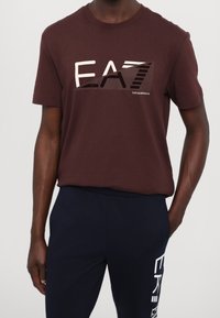 Tummanpunainen, puuvillainen T-paita, jossa rohkea graafinen "EA7"-logo kontrastiväreissä. Pyöreä pääntie ja lyhyet hihat.