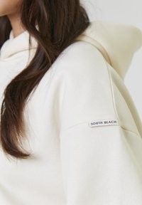 Vrouw met lang donker haar die een crèmekleurige hoodie draagt met een "SOUTH BEACH" label op de bovenarm nabij de schouder.
