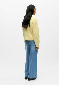 Vaaleankeltainen neulepaita, jossa on teksturoitu kuvio, yhdistettynä leveälahkeisiin sinisiin farkkuihin. Asu on rento ja siinä on resorireunukset hihoissa.