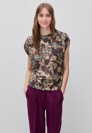 Junge Frau mit langen braunen Haaren, die ein kurzärmeliges braunes Blumenoberteil und violette Hose trägt, mit Händen in den Taschen, vor einem schlichten Hintergrund stehend.
