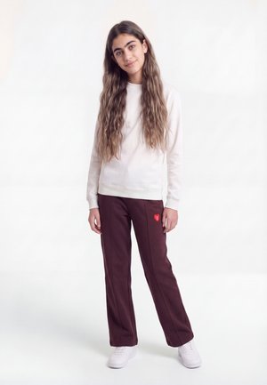 Jeune femme aux longs cheveux ondulés portant un sweat-shirt blanc, un pantalon bordeaux avec un petit détail cœur rouge et des baskets blanches, debout devant un fond blanc.
