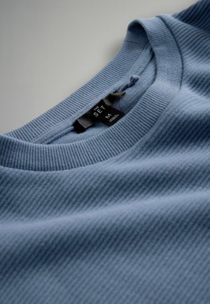 Maglietta a costine color azzurro chiaro con scollo tondo e texture morbida. Presenta un'etichetta in tessuto nero con informazioni su marca e taglia.