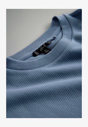 Maglietta a costine color azzurro chiaro con scollo tondo e texture morbida. Presenta un'etichetta in tessuto nero con informazioni su marca e taglia.