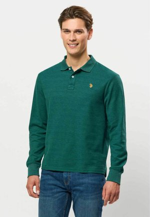 Jeune homme souriant portant un polo vert à manches longues et un jean bleu, debout devant un fond clair uni.