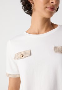 T-shirt bianca a maniche corte con alette della tasca in beige a motivo houndstooth texturizzato e dettagli di bottoni dorati sul davanti. Finitura in tessuto liscia.