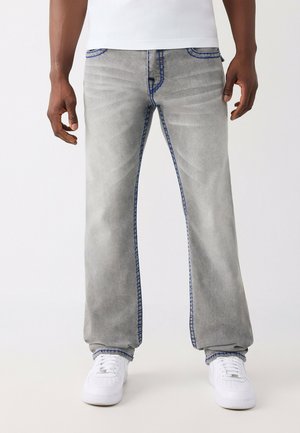 Een persoon draagt lichtgrijze straight-leg jeans met blauwe stiksels en witte sneakers, staand tegen een witte achtergrond.