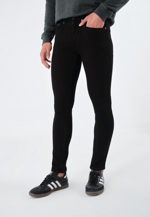 Mann trägt schlanke schwarze Jeans, dunkelgraues Langarmshirt und schwarze Adidas-Sneaker mit weißen Streifen vor weißem Hintergrund.