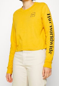 Gul långärmad cropped topp i bomull, med en fyrkantig logotyp på bröstet och "Huf worldwide" tryckt i svart längs ärmen.