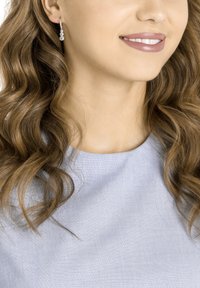 Silver earrings with three cascading clear gemstones, worn by a person with wavy hair, against a light blue fabric background.