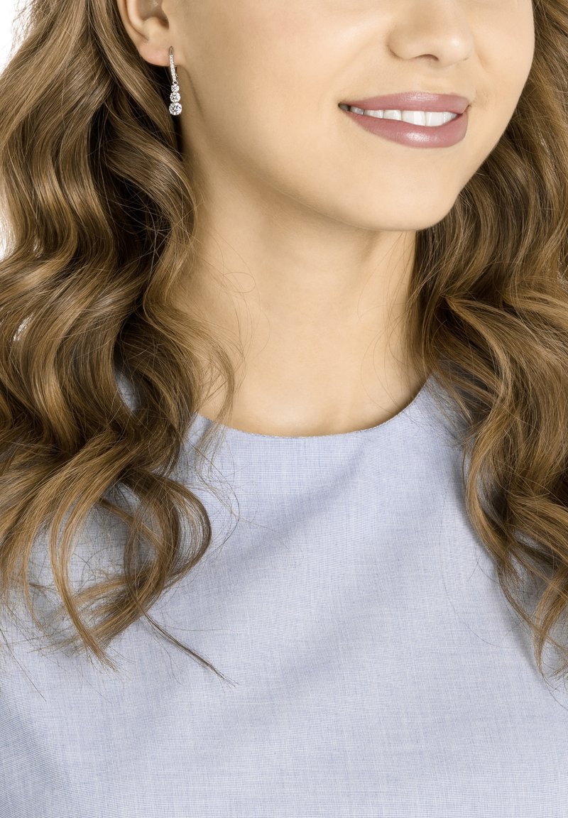 Silver earrings with three cascading clear gemstones, worn by a person with wavy hair, against a light blue fabric background.