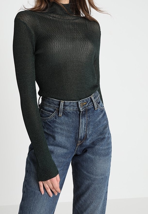 Femme portant un col roulé côtelé à manches longues ajusté de couleur vert foncé et un jean en denim bleu taille haute.