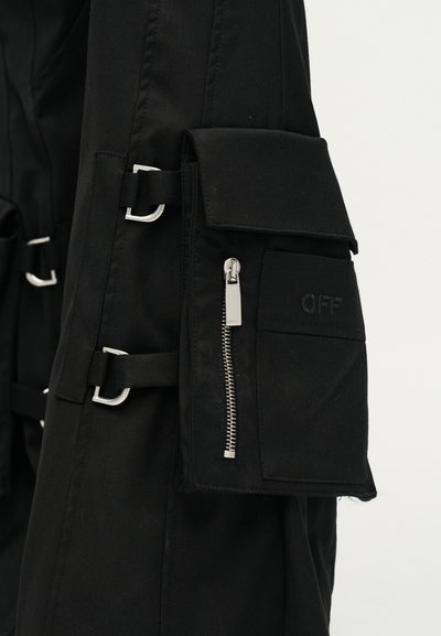 Μαύρο υφασμάτινο ένδυμα με πολλαπλές ασημένιες αγκράφες και μια τσέπη με φερμουάρ που διαθέτει πτερύγιο με την ετικέτα "OFF" στο πλάι.