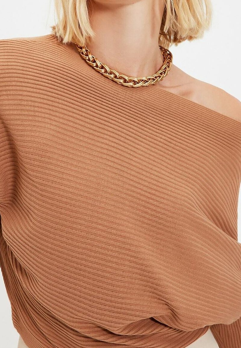Femme blonde portant un pull côtelé camel à épaules dénudées et un collier chaîne en or épais.