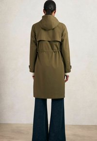 Manteau imperméable vert olive avec capuche, taille cintrée et détail de rabat au dos, associé à un pantalon en denim foncé à jambe large.