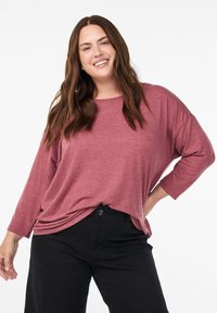 Smiling woman with long brown hair wears a loose mauve long-sleeve top and black pants, posing against a white background.