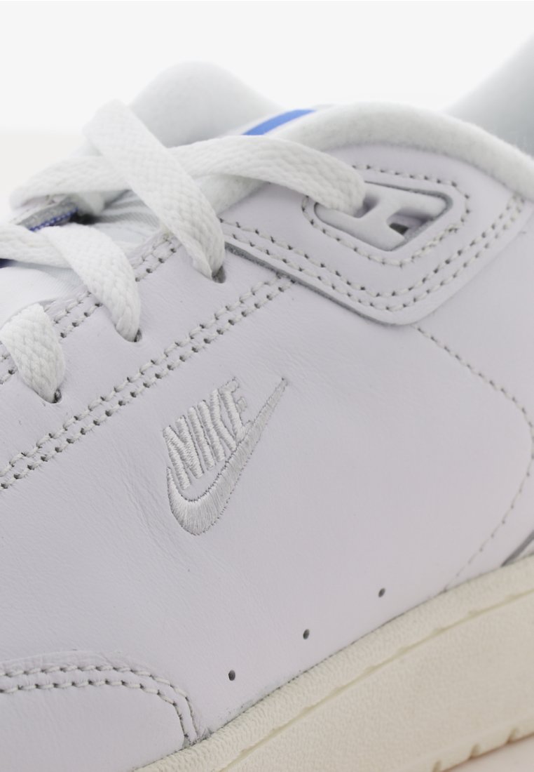Weiße Leder-Sneaker mit dezentem Nike-Logo. Besitzt weiße Schnürsenkel, eine glatte Oberseite und eine beiges Gummisohle. Blauer Akzent nahe der Zunge.