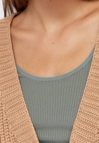 Beżowy sweter z dzianiny z szerokim otwartym przodem, nałożony na jasnoniebieski top z żeberkami. Skupienie na fakturze i kontraście kolorów.