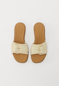 Sandaler med bred såle og en kremfarget syntetisk overdel, dekorert med en stor firkantet spenne, plassert på en teksturert brun såle med flatt design.