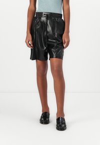 Zwarte leren shorts met een gladde textuur, elastische tailleband en een koorddetail, gecombineerd met zwarte loafers en een lichtblauw top.
