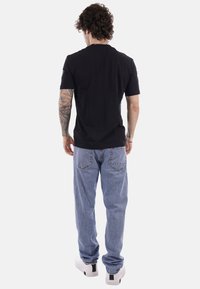 Uomo con capelli ricci che indossa una maglietta nera, jeans azzurri, sneakers bianche e tatuaggi visibili sulle braccia, in piedi di spalle.