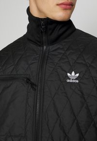 Svart vadderad jacka med hög krage, framdragkedja och en liten ficka med dragkedja på bröstet. Adidas-logotypen syns på vänster sida.