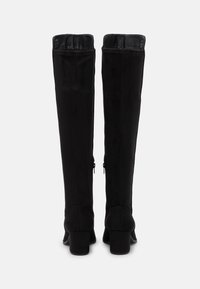 Pár vysokých černých semišových bot na podpatku s blokovými podpatky, se zipem na zadní straně, zobrazené zezadu na bílém pozadí.