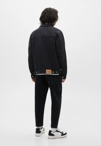 Personne aux cheveux noirs mi-longs vue de dos, portant une veste et un pantalon en jean noir avec des baskets blanches et noires sur un fond blanc.