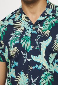 Košile s krátkým rukávem s námořnickým pozadím a živými vzory tropických listů v zelené a světle modré barvě, doplněná světle hnědými knoflíky.