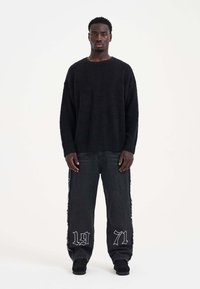 Zwarte fluffy trui met lange mouwen, gecombineerd met zwarte denim jeans waarop het jaar "1971" in het wit op de broekspijpen is geborduurd.