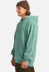 Turquoise hoodie made of cotton with a front pocket and a drawstring hood, paired with brown knit beanie. Simple, casual design.