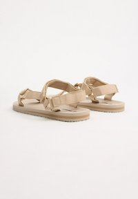 Sandalias beige con correas de tela, hebillas ajustables y suelas de goma planas con un patrón texturizado para mayor agarre. Diseño minimalista.