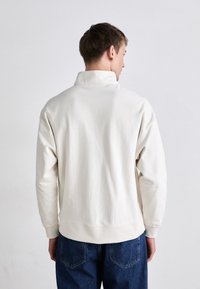 Suéter crema claro con cuello alto, mangas largas y dobladillo acanalado. Lleva puesto unos vaqueros de mezclilla azul. Tejido suave con textura mínima.