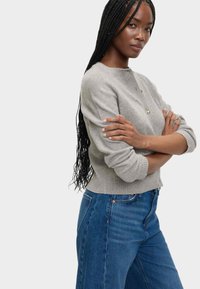 Grå stickad tröja med rund halsringning och tre dekorativa knappar, parat med högmidjade blå jeans. Texturen verkar mjuk.