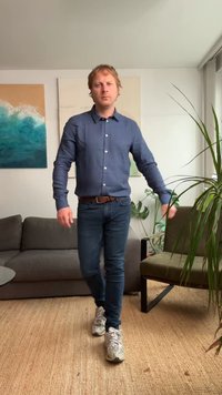 Zila pogu krekls, tumšie džinsi, pelēki sporta apavi. Vīrietis staigā pa austamajam paklājam ar pelēku dīvānu un zaļu augu labi apgaismotā interjerā.