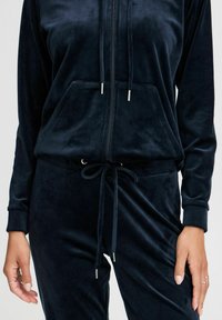 Mörkblå velourträningsoverall med zip-up hoodie och stor framficka. Har dragsko i midjan och silverdetaljer. Mjuk textur.