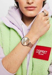 Runde silberne Uhr mit einem gebürsteten roségoldenen Zifferblatt, Metall-Mesh-Armband, am Handgelenk getragen, das gegen eine grüne Fleece-Weste mit einem roten Logopatch ist.