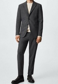 Costume gris en tissu texturé, comprenant un blazer à un bouton, un pantalon assorti et des bottines noires. Pull crème uni en dessous.