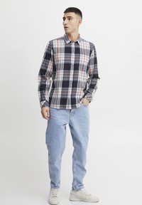 Camisa de cuadros en tonos azul marino, blanco y naranja; de manga larga, con botones en la parte delantera, de material de algodón. Combinada con vaqueros de corte claro y zapatillas blancas.